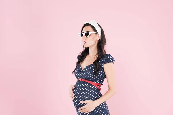 Беременная женщина в солнечных очках жует жвачку, изолированную от розового — стоковое фото