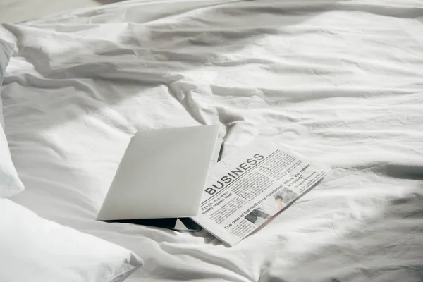 Laptop near business newspaper on white bedding in bedroom — Stock Photo