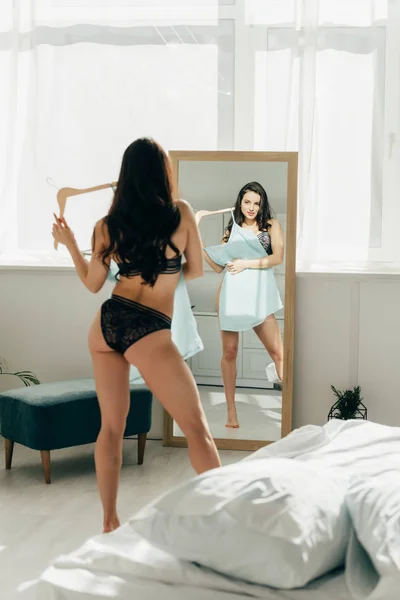 Attraktive Frau hält Kleiderbügel mit blauem Kleid, während sie in Spitzenunterwäsche steht und in den Spiegel schaut — Stockfoto