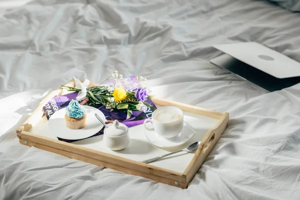 Ноутбук рядом с деревянным подносом с вкусным завтраком на кровати — стоковое фото