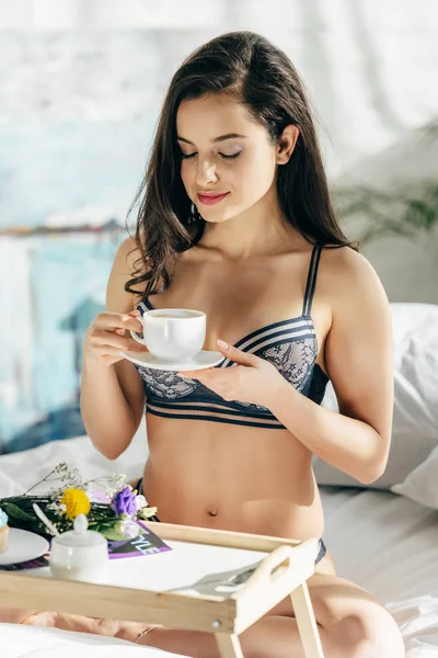 Hermosa mujer morena en ropa interior sosteniendo taza mientras está sentado cerca de bandeja de madera con desayuno - foto de stock