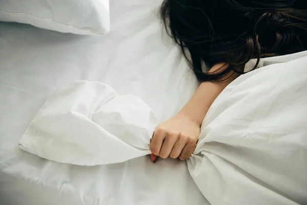 Vista recortada de la mujer sosteniendo ropa de cama blanca en la mano mientras está acostado en la cama - foto de stock