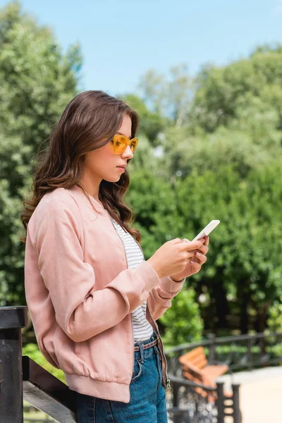 Attractive girl in sunglasses using smartphone while standing in park — Stock Photo