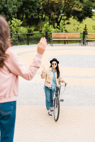 Back view of girl waving hand near cheerful friend with bicycle — Stock Photo