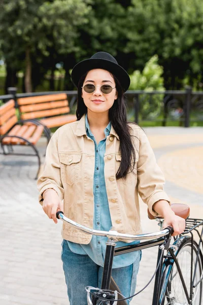 Hübsches Mädchen mit Sonnenbrille und Hut steht mit Fahrrad im Park — Stockfoto