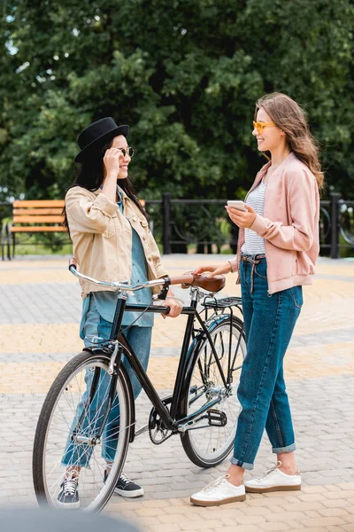 Cheerful girls in sunglasses smiling while standing near bike in park — Stock Photo