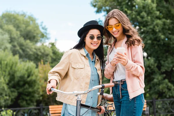 Cheerful girls in sunglasses smiling while looking at smartphone and standing near bike in park — Stock Photo