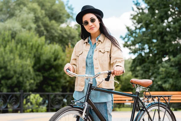 Fille heureuse dans les lunettes de soleil et chapeau debout avec vélo dans le parc — Photo de stock