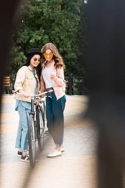 Enfoque selectivo de chicas alegres en gafas de sol sonriendo mientras mira el teléfono inteligente y de pie cerca de la bicicleta en el parque - foto de stock
