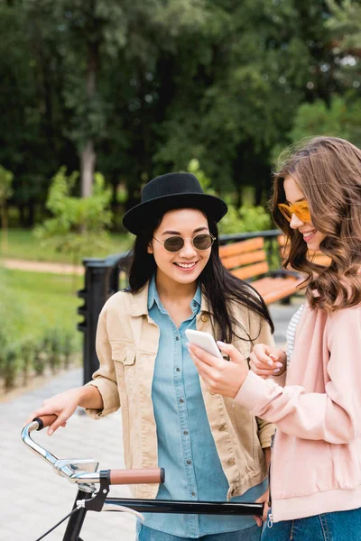 Счастливые девушки в солнечных очках улыбаются, глядя на смартфон и стоя возле велосипеда в парке — стоковое фото