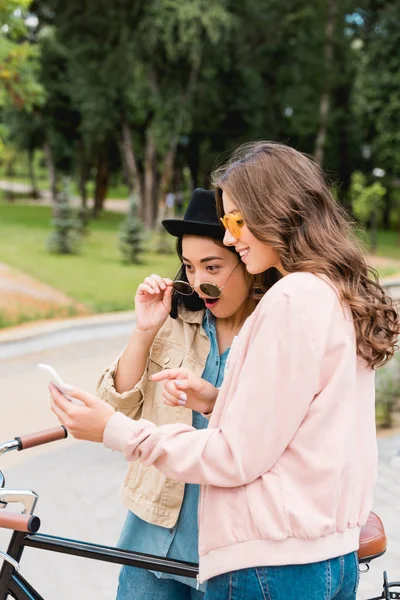 Überraschtes Mädchen mit Sonnenbrille und Hut blickt auf Smartphone und steht neben hübscher Freundin im Park — Stockfoto