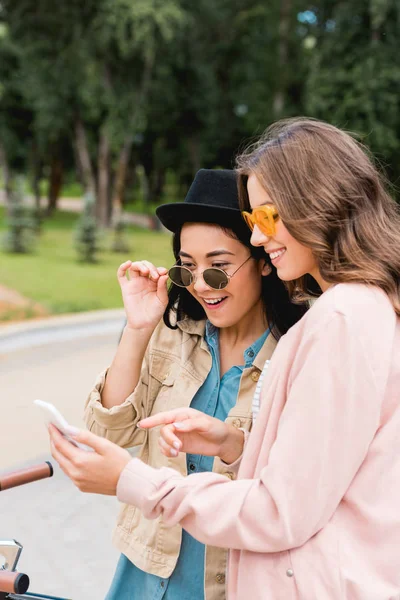 Chicas atractivas en gafas de sol sonriendo mientras mira el teléfono inteligente y de pie cerca de la bicicleta en el parque - foto de stock