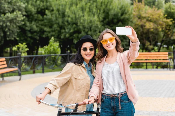 Meninas atraentes em óculos de sol sorrindo ao tomar selfie no parque — Fotografia de Stock