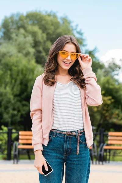 Pretty girl smiling while touching sunglasses and holding smartphone — Stock Photo