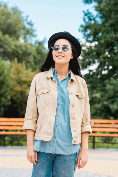 Счастливая девушка в солнцезащитных очках и шляпе улыбается, стоя в парке — стоковое фото