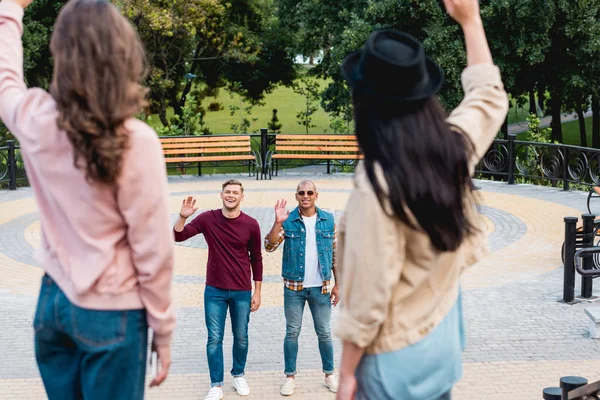 Foyer sélectif de jeunes hommes multiculturels joyeux agitant la main et souriant à des amies dans le parc — Photo de stock