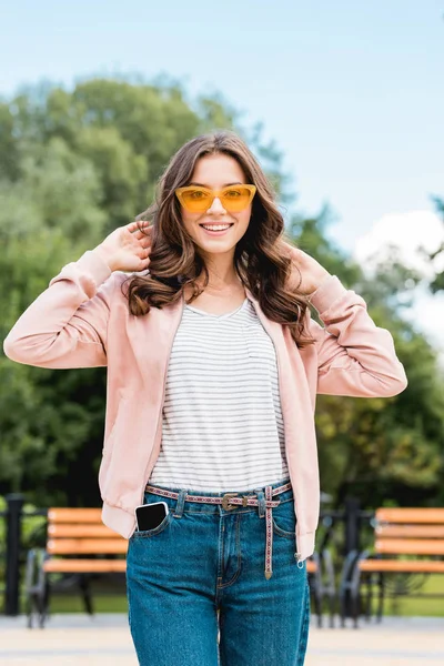 Bonita chica en gafas de sol sonriendo mientras se toca el pelo en el parque - foto de stock