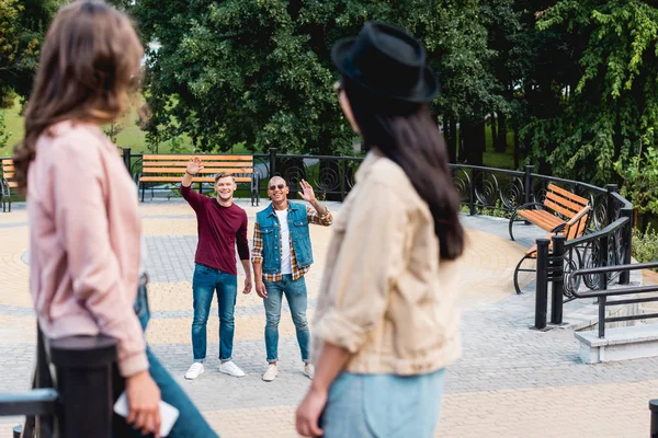 Selective focus of happy young men waving hand and smiling to female friends in park — Stock Photo
