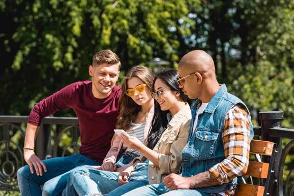 Cheerful multicultural friends sitting on bench and looking at smartphone — Stock Photo