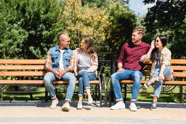 Amis multiculturels joyeux assis sur des bancs dans le parc — Photo de stock