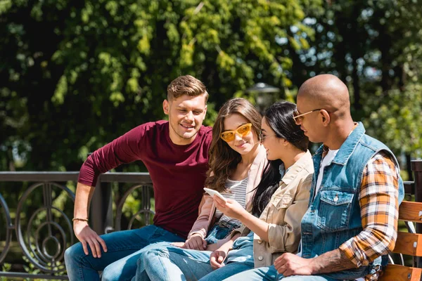 Amis multiculturels joyeux assis sur le banc et regardant fille tenant smartphone — Photo de stock