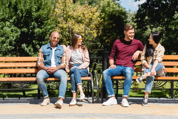 Amigos multiculturais alegres sorrindo enquanto sentados em bancos no parque — Fotografia de Stock