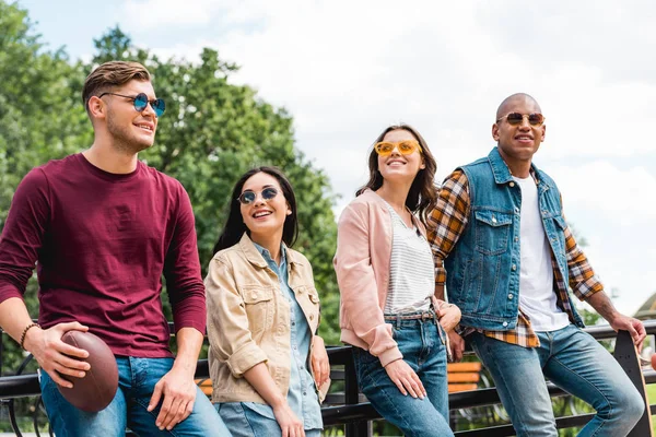 Joyeux jeunes hommes multiculturels dans des lunettes de soleil debout avec longboard et football américain près de jolies filles — Photo de stock