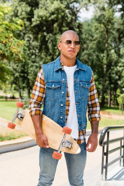Handsome african american man in sunglasses standing with longboard — Stock Photo