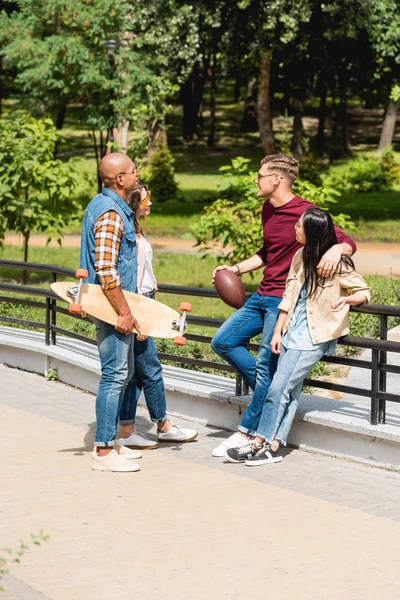 Feliz multicultural jóvenes en gafas de sol de pie con longboard y fútbol americano cerca de chicas bonitas - foto de stock