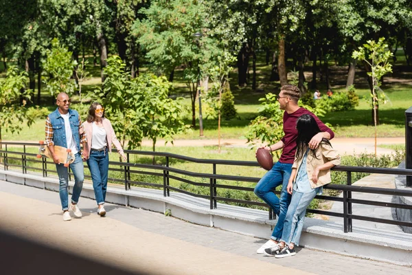 Selektiver Fokus des glücklichen afrikanisch-amerikanischen Mannes, der Longboard hält und mit Mädchen in der Nähe von Freunden spazieren geht — Stockfoto