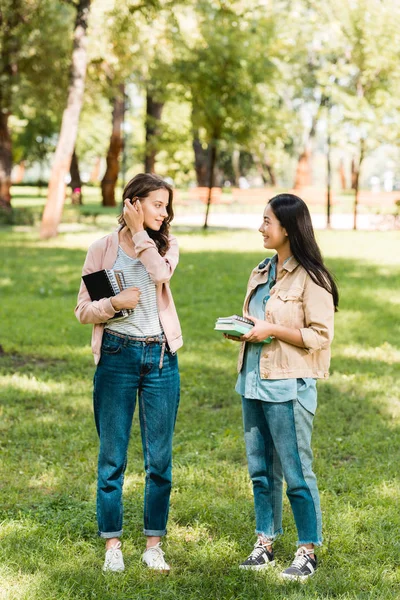Attractive girls holding books and looking at each other while standing in park — Stock Photo