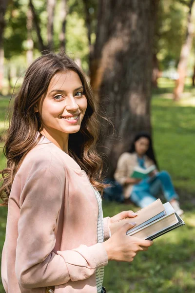 Selective focus of cheerful girl holding books with friend on background — Stock Photo