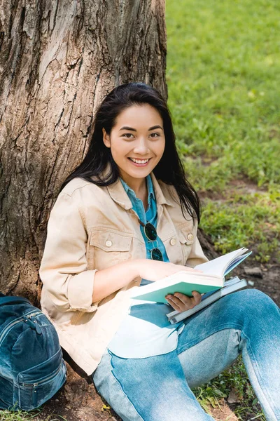 Bastante estudiante sonriendo mientras está sentado en el parque con libros - foto de stock