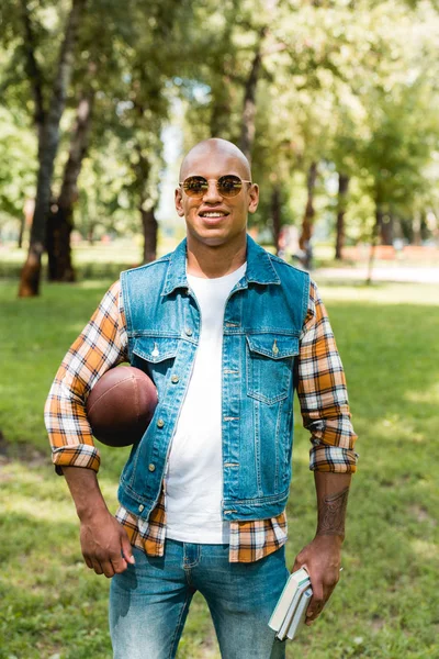 Alegre afroamericano hombre en gafas de sol celebración de libros y fútbol americano - foto de stock
