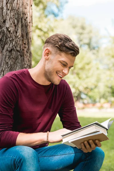 Alegre estudiante leyendo libro y sonriendo mientras está sentado en el parque - foto de stock