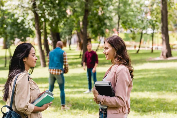 Selective focus of cheerful girls talking while standing with books near students playing american football — Stock Photo