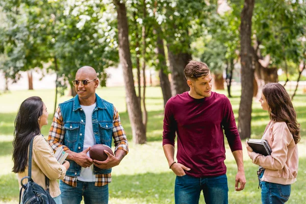 Fröhliche multikulturelle Studenten unterhalten sich, während sie mit Büchern im Park stehen — Stockfoto