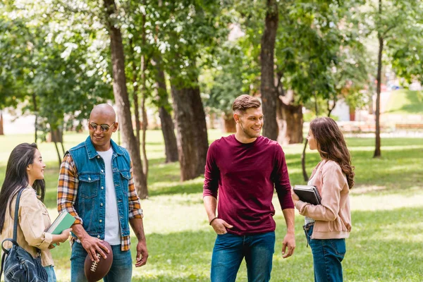 Chicas atractivas hablando con estudiantes multiculturales alegres mientras están de pie con libros en el parque - foto de stock