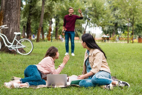 Selective focus of girls sitting on blanket near laptop and waving hand near young man — Stock Photo