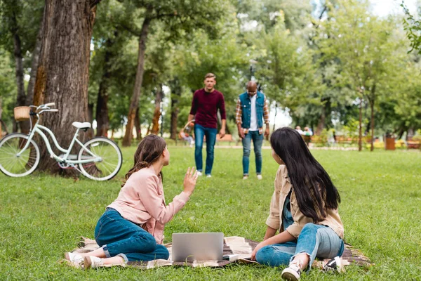 Selective focus of girls sitting on blanket near laptop and waving hand near multicultural men — Stock Photo