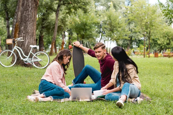 Handsome man holding longboard and sitting with girls on blanket near laptop — Stock Photo