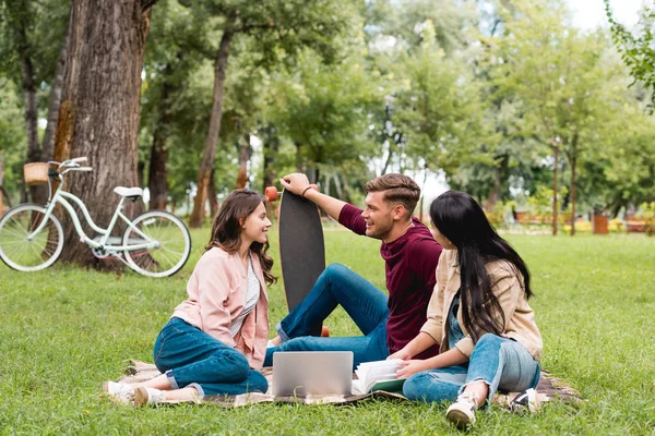 Handsome man holding longboard and sitting with cheerful girls on blanket near laptop — Stock Photo