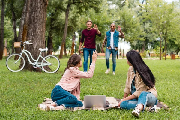 Messa a fuoco selettiva di ragazze sedute su coperta vicino al computer portatile e guardando allegri uomini multiculturali che salutano le mani — Foto stock