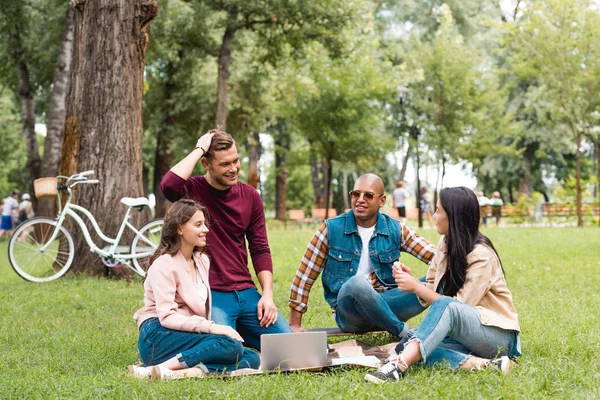 Cheerful multicultural men sitting with attractive girls on blanket near laptop — Stock Photo