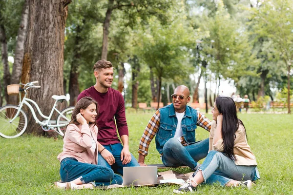 Hombres multiculturales felices sentados con chicas atractivas en manta cerca de la computadora portátil - foto de stock