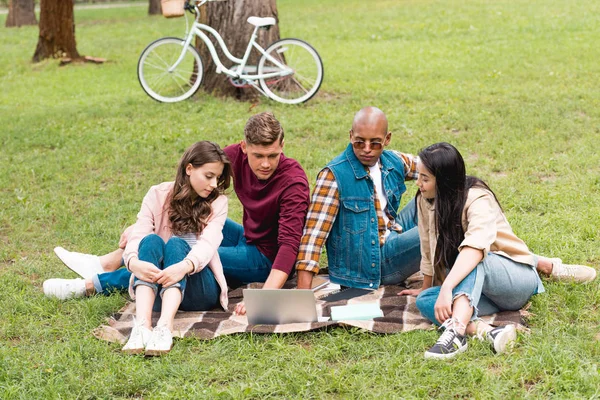 Beaux jeunes hommes multiculturels assis avec des filles attrayantes sur couverture près d'un ordinateur portable — Stock Photo