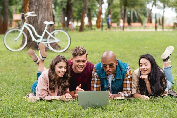 Feliz multicultural jóvenes que yacen con hermosas chicas en la manta y mirando a la computadora portátil - foto de stock