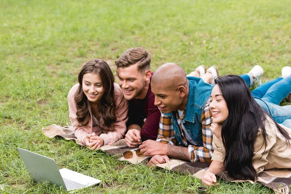 Happy multicultural friends lying on blanket and looking at laptop in park — Stock Photo