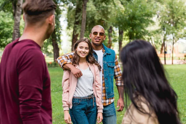 Foco seletivo do homem americano africano abraçando menina enquanto está com os amigos no parque — Fotografia de Stock