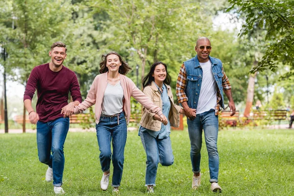 Cheerful multicultural friends holding hands and running in park — Stock Photo